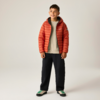 Junge steht vor neutralem Hintergrund, trägt orange Steppjacke, beigefarbenen Pullover, schwarze Hosen und weiße Sneakers mit grünen Schnürsenkeln