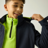 Junge lächelt, trägt hellgrünen Fleece und dunkle Jacke, zieht am Kragen, heller Hintergrund
