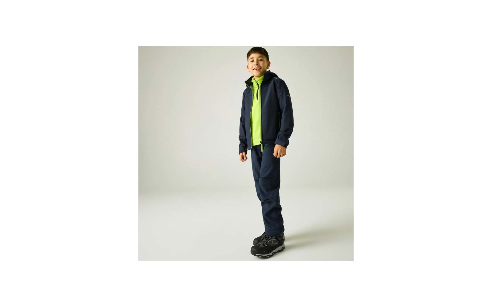 Ein Junge steht vor hellem Hintergrund, trägt eine dunkelblaue Kapuzenjacke, ein neongrünes Zip-up darunter, dunkelblaue Hose und schwarze Sportschuhe
