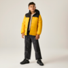 Junge steht vor hellem grauem Hintergrund, trägt gelbe Steppjacke mit schwarzem Kragen, helles T‑Shirt, weite schwarze Jeans mit weißen Nähten und schwarze Sneakers; Hände an den Seiten, Blick nach oben rechts