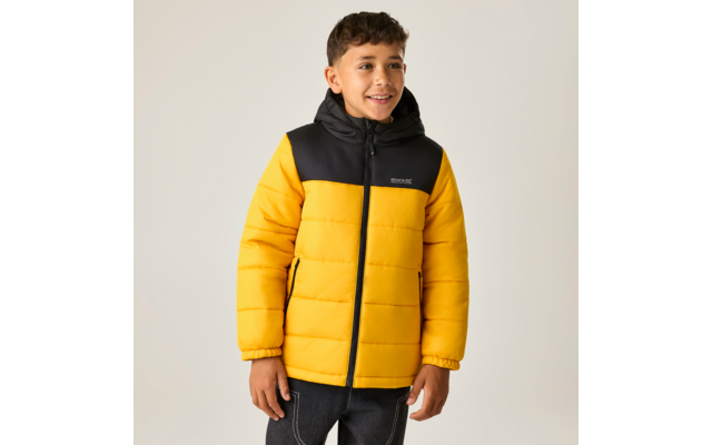 Junge steht vor hellem Hintergrund, trägt gelb-schwarze gesteppte Kapuzenjacke und dunkle Jeans