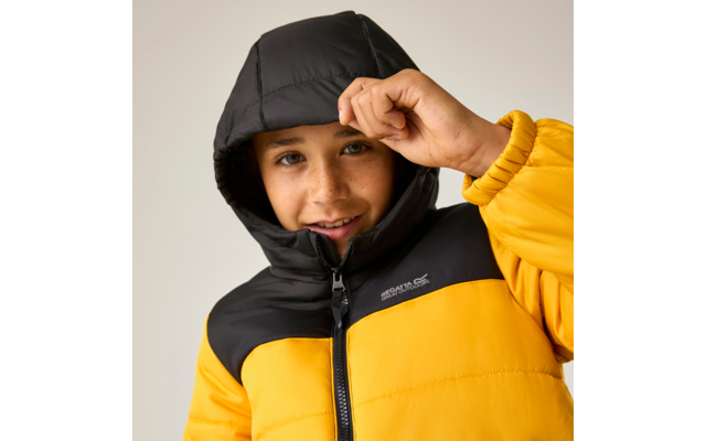 Kind in gelber und schwarzer Steppjacke mit Kapuze, Hand an der Kapuze, heller einfarbiger Hintergrund