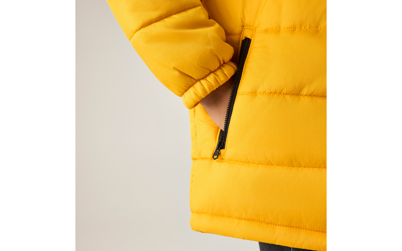 Hand in Reißverschlusstasche einer gelben Steppjacke, sichtbarer Ärmel und Saum vor hellem Hintergrund