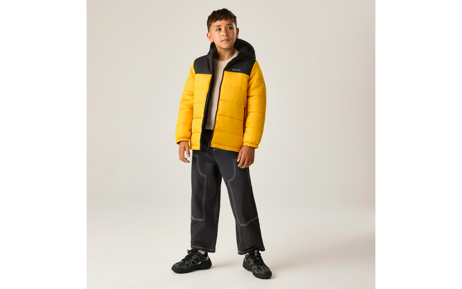 Junge steht vor hellem grauem Hintergrund, trägt gelbe Steppjacke mit schwarzem Kragen, helles T‑Shirt, weite schwarze Jeans mit weißen Nähten und schwarze Sneakers; Hände an den Seiten, Blick nach oben rechts
