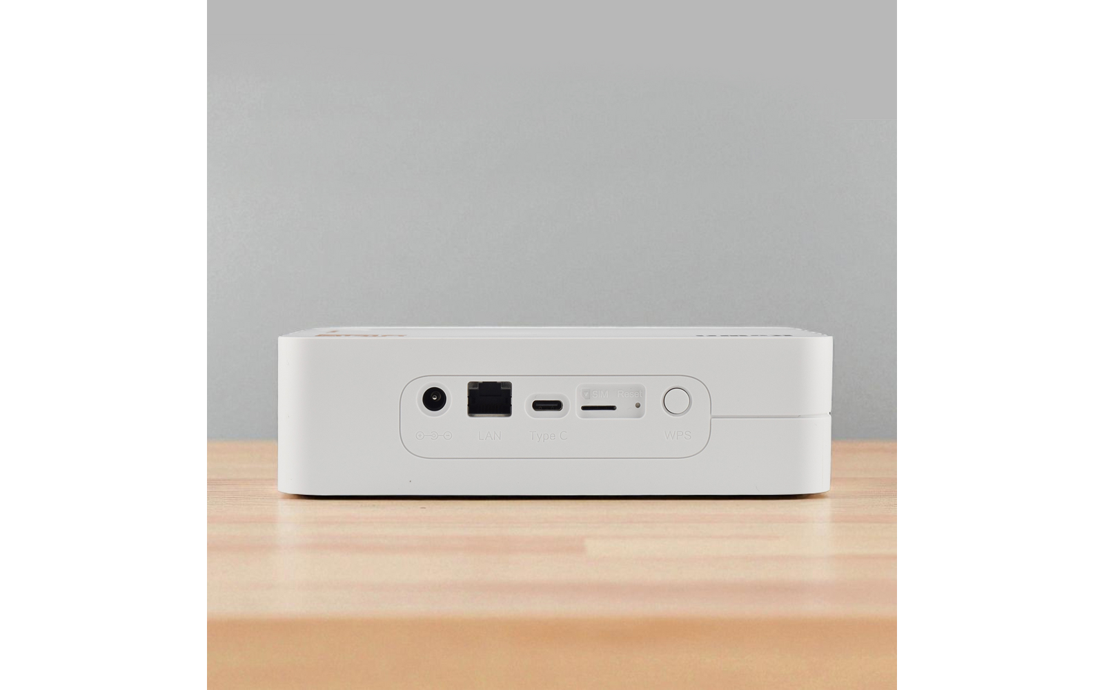 Weißes rechteckiges Gerät mit runder Netzanschlussbuchse, Ethernetbuchse, USB‑C‑Anschluss, Kartenschlitz und rundem Knopf, auf Holzoberfläche vor grauer Wand
