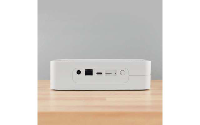 Weißes rechteckiges Gerät mit runder Netzanschlussbuchse, Ethernetbuchse, USB‑C‑Anschluss, Kartenschlitz und rundem Knopf, auf Holzoberfläche vor grauer Wand
