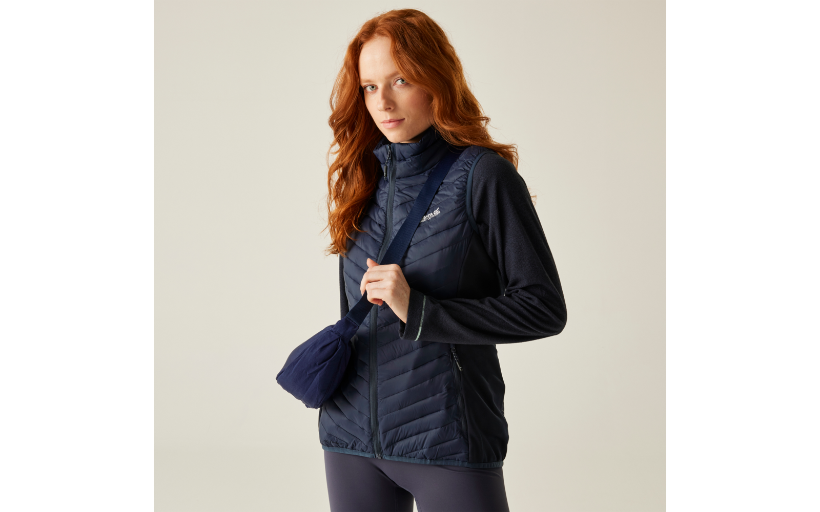 Vrouw met rood haar draagt een navy doorgestikte bodywarmer, donker shirt, donkere legging en een klein schoudertasje voor een lichte achtergrond