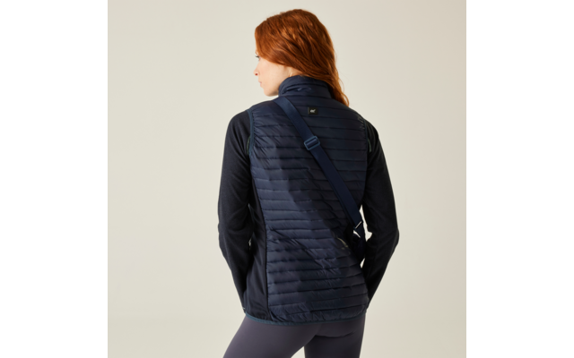Persoon met rood haar van achteren gezien, draagt een marineblauwe, gewatteerde bodywarmer, donker shirt met lange mouwen en een diagonale tasriem, lichte achtergrond