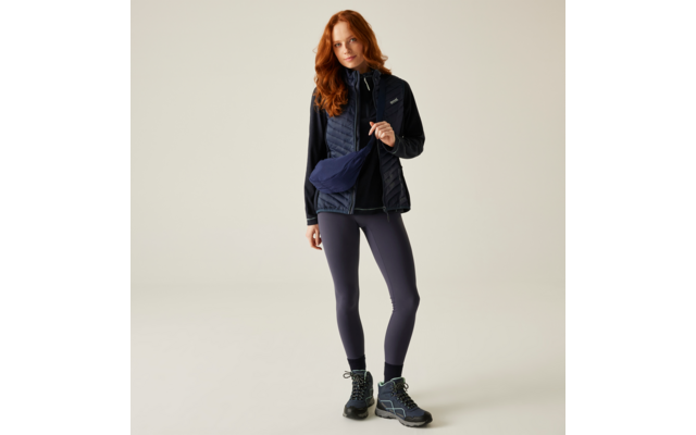 Roodharige vrouw staand in outdoorkleding tegen een lichte achtergrond, draagt een doorgestikte bodywarmer, jas, legging, wandelschoenen en een crossbodytas