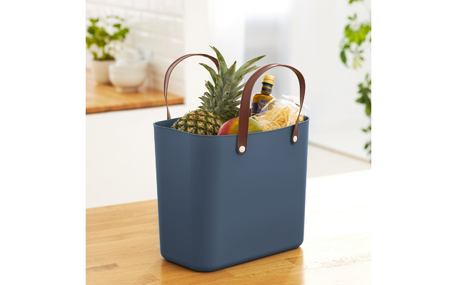 Blauer Aufbewahrungskorb mit Ledergriffen auf Holzoberfläche, darin eine Ananas und eine Vase mit Pflanze