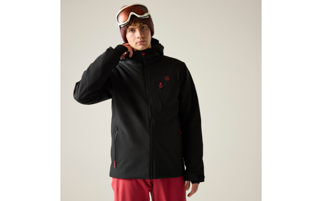 Person in schwarzer Jacke mit roten Reißverschlussdetails, roter Hose, brauner Strickmütze und Skibrille auf der Stirn, eine Hand am Jackenkragen, einfarbiger heller Hintergrund