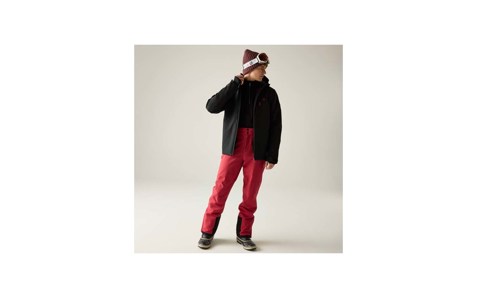 Person in schwarzer Jacke, roter Hose, Mütze und Skibrille, steht und blickt zur Seite