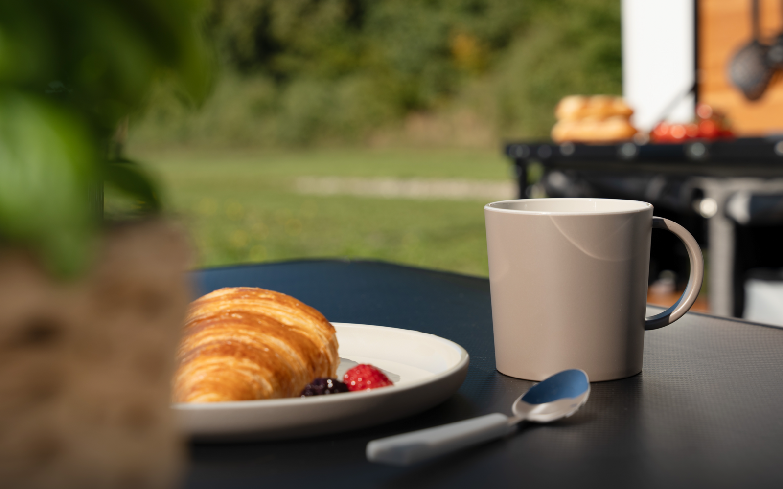 Auf einem schwarzen Tisch: ein Teller mit Croissant und roten sowie dunklen Beeren, daneben ein beigefarbener Becher und ein Löffel; im Vordergrund unscharfe Pflanze, im Hintergrund unscharfes schwarzes Gerät mit Brötchen und Rasen