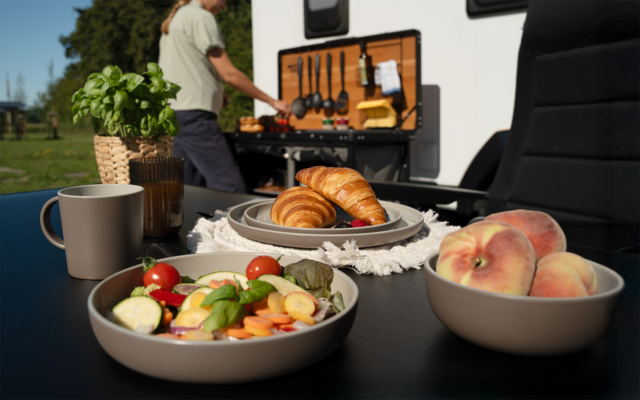 Tisch im Freien mit Schüssel Salat, Teller mit Croissants, Schale mit Pfirsichen, Kaffeetasse und Kräutertopf; im Hintergrund offene Wohnmobilküche und eine Person