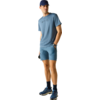 Mann in blauem Sportoutfit mit Kappe, T‑Shirt, Shorts, dunklen Socken und Sportschuhen, steht und blickt nach rechts, eine Hand in der Hosentasche