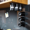 Drei schwarze zylindrische Dosen mit weißen Etiketten unter einer Holzschrankkante; rechts drei schwarze runde Magnetdosen an einer vertikalen Wandhalterung; links ein hängendes Papierkarte mit gelbem Aufkleber; dunkle glänzende Rückwand mit Spiegelu