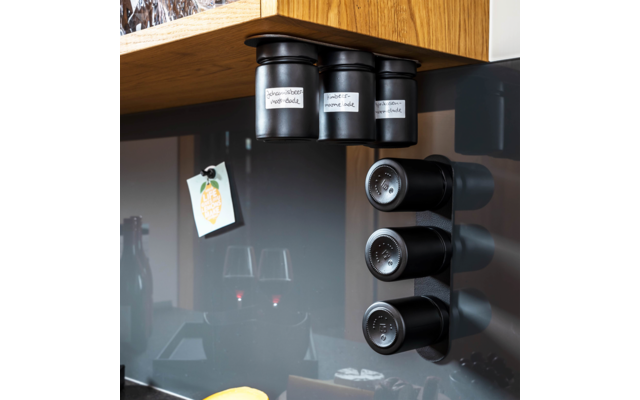 Drei schwarze zylindrische Dosen mit weißen Etiketten unter einer Holzschrankkante; rechts drei schwarze runde Magnetdosen an einer vertikalen Wandhalterung; links ein hängendes Papierkarte mit gelbem Aufkleber; dunkle glänzende Rückwand mit Spiegelu
