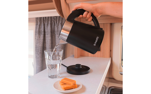 Hand houdt zwarte waterkoker en giet water in een glas op een aanrecht; kokerbasis met snoer, klein wit bordje met drie gele plakken, houten kast en gordijn op de achtergrond