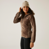 Frau trägt eine braune Jacke, schwarze Hose und eine beige Mütze, steht vor hellem Hintergrund