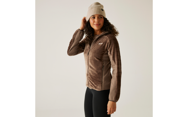 Frau trägt eine braune Jacke, schwarze Hose und eine beige Mütze, steht vor hellem Hintergrund
