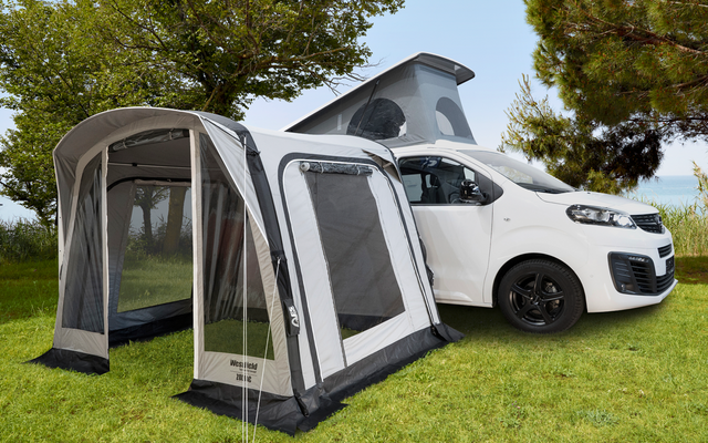 Witte camper met open daktent en aangesloten grijze voortent, staand op groen gras onder bomen