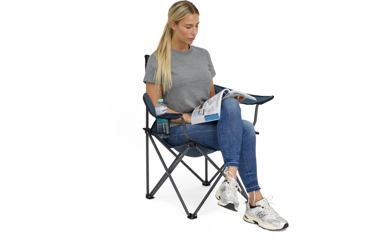 Frau mit langen blonden Haaren sitzt in blauem Klappstuhl und liest ein Magazin; trägt graues T‑Shirt, blaue Jeans und weiße Sneakers; Wasserflasche im Getränkehalter an der Armlehne