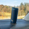 Gobelet de voyage bleu avec inscription blanche posé dans une cavité ronde sur le tableau de bord d'une voiture ; en arrière-plan, route et arbres flous visibles à travers le pare-brise