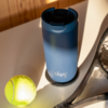 Gobelet bleu avec couvercle et logo silwy sur une table, à côté d'une balle de tennis et d'une raquette, lumière du soleil et ombres
