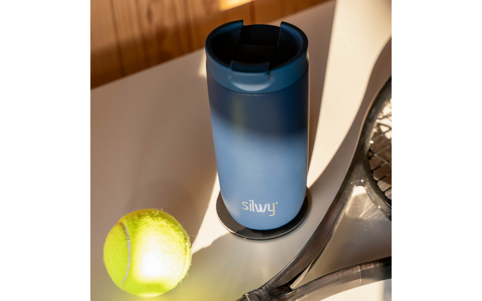Gobelet bleu avec couvercle et logo silwy sur une table, à côté d'une balle de tennis et d'une raquette, lumière du soleil et ombres