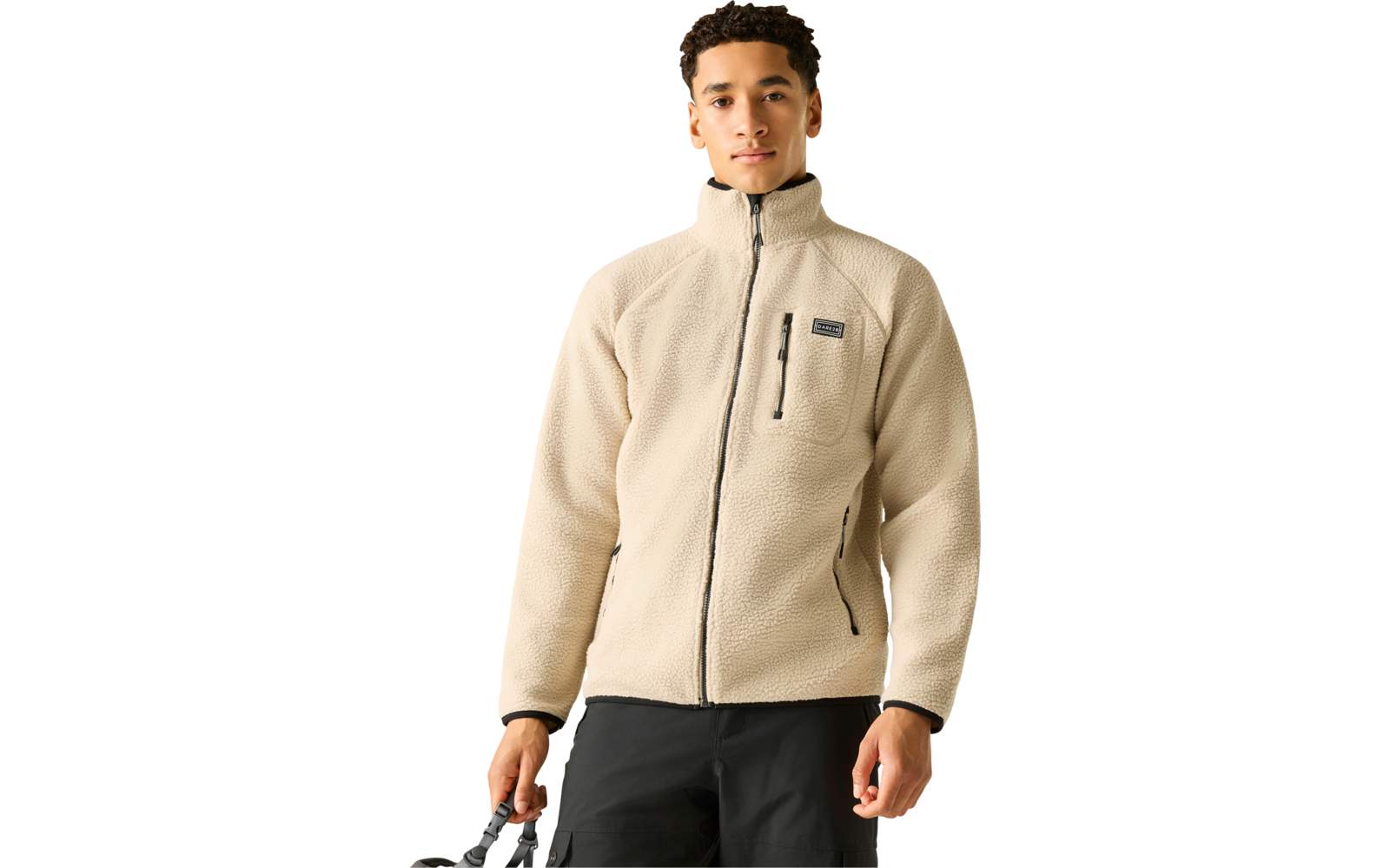 Person mit beige Fleecejacke (Vollreißverschluss, vertikale Brusttasche, seitliche Reißverschlusstaschen), schwarze Hose, hält schwarzen Taschenhenkel, weißer Hintergrund