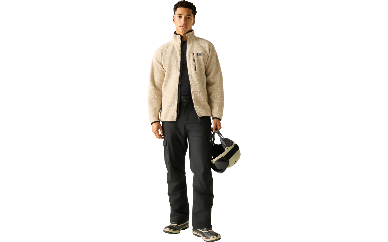 Person steht vor hellem Hintergrund, trägt beige Fleecejacke, schwarzes Oberteil, schwarze Hose und schwarze Schuhe, hält einen Motorradhelm in der rechten Hand