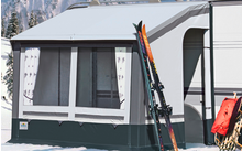 Wohnwagen mit angebautem Vorzelt auf verschneiter Fläche, Skier und Stöcke lehnen am Vorzelt, im Hintergrund schneebedeckte Tannen und Berge