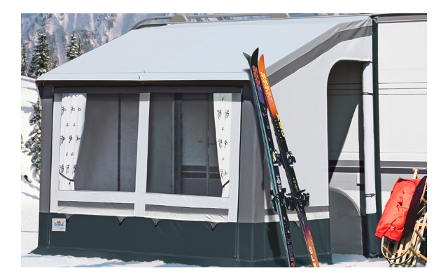 Wohnwagen mit angebautem Vorzelt auf verschneiter Fläche, Skier und Stöcke lehnen am Vorzelt, im Hintergrund schneebedeckte Tannen und Berge