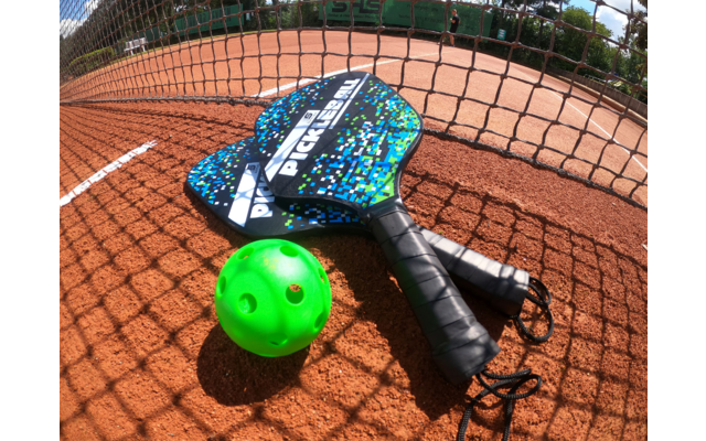 Zwei Padel-Schläger mit blau-weißem Muster liegen auf einem Tennisplatz, davor liegt ein grüner Ball