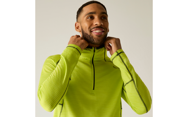 Personne aux cheveux courts et foncés et barbe, souriante, porte un haut de sport vert néon à fermeture éclair, tient le col des deux mains, fond clair