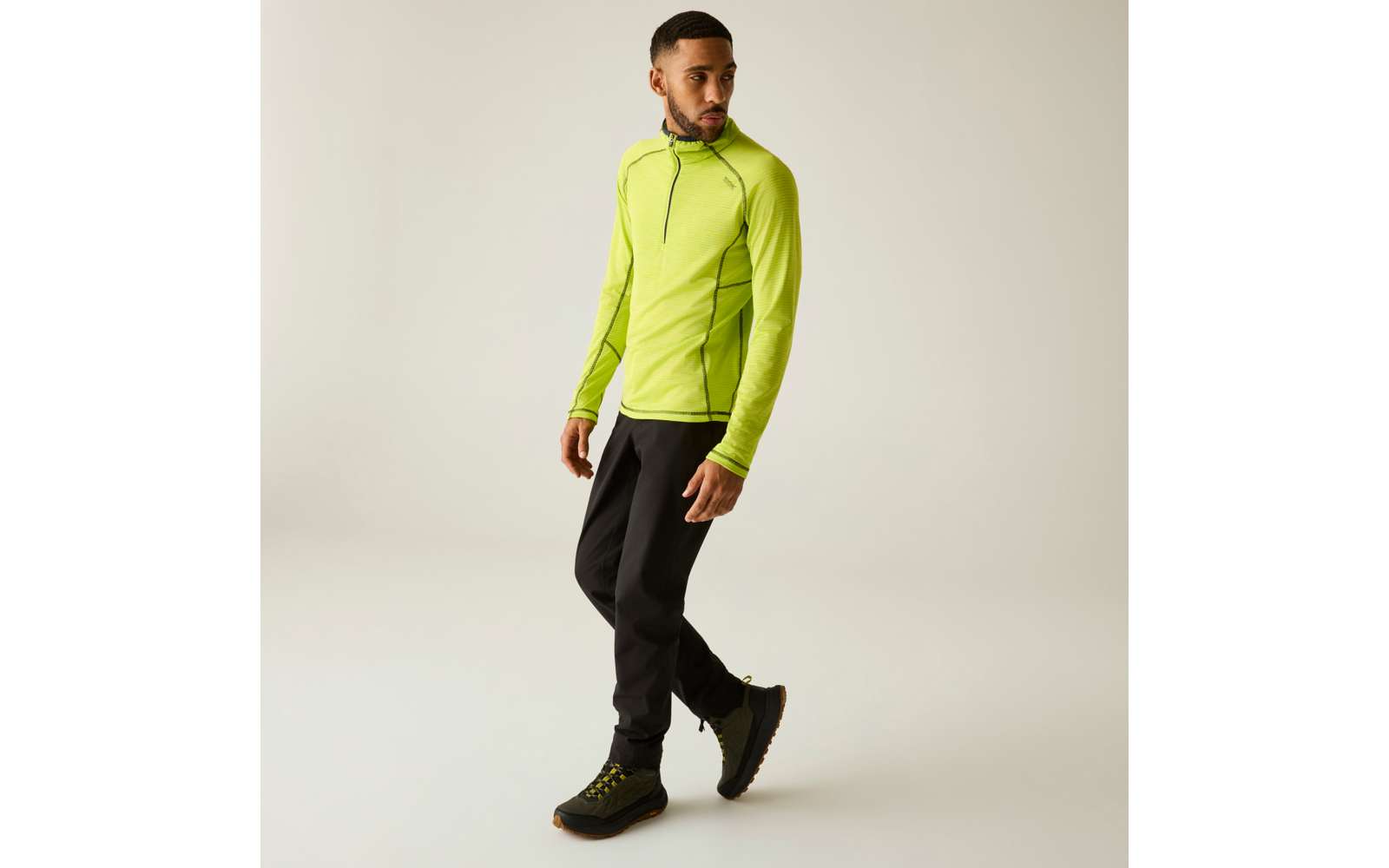 Homme en haut à manches longues vert néon avec petit zip, pantalon noir et chaussures de randonnée noires, marchant et regardant sur le côté sur fond clair