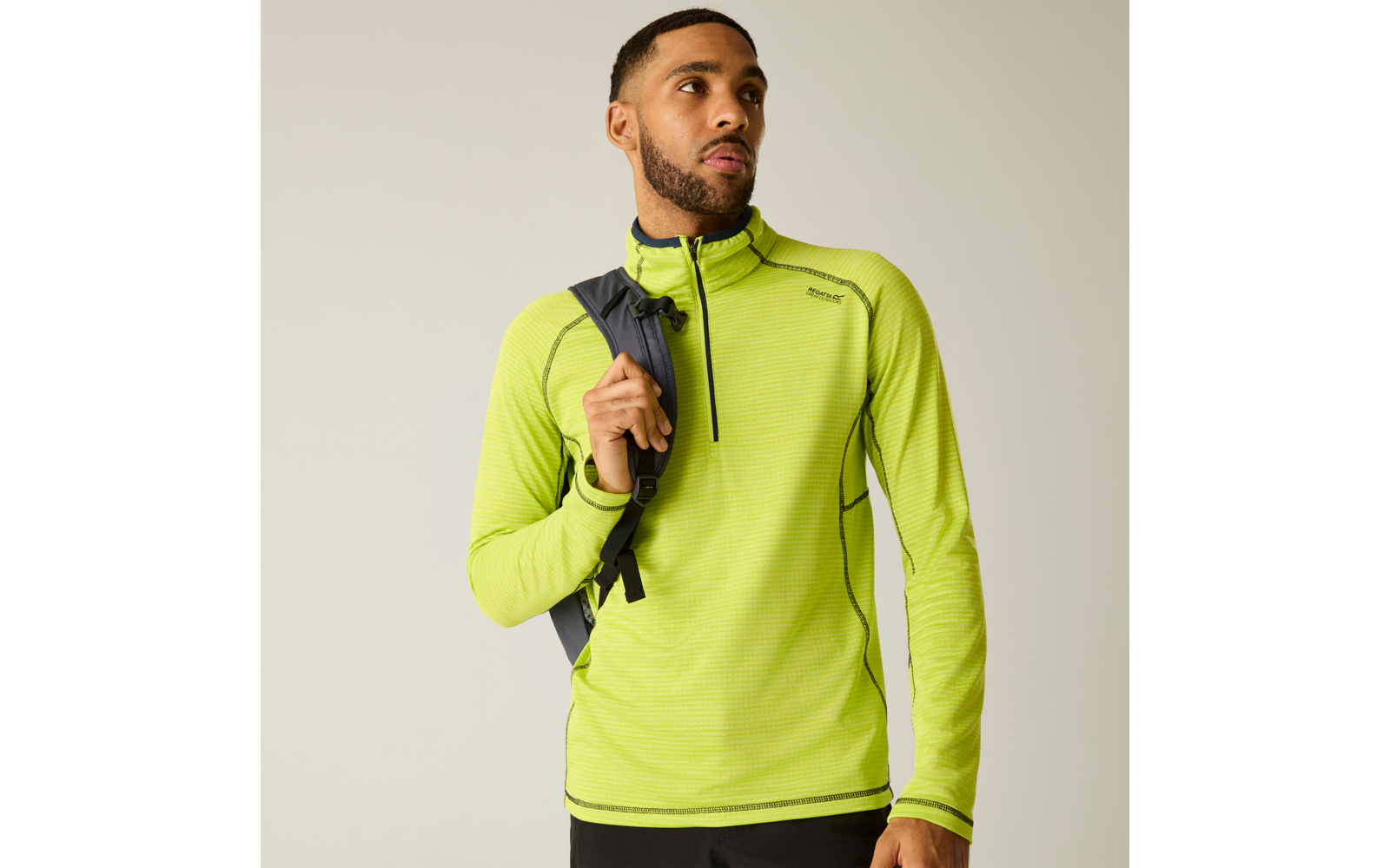 Personne devant un fond uni clair, portant un haut à manches longues vert néon avec une courte fermeture éclair au col, un sac à dos noir sur une épaule et un pantalon noir, regardant légèrement vers la droite