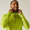 Personne aux cheveux courts et foncés et barbe, souriante, porte un haut de sport vert néon à fermeture éclair, tient le col des deux mains, fond clair