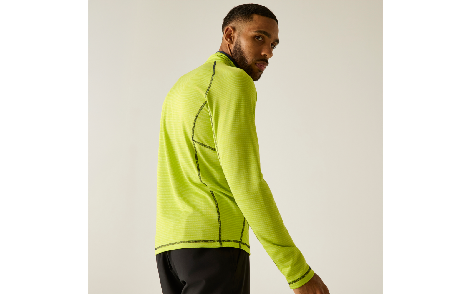 Personne portant un haut de sport vert fluo à manches longues et un pantalon noir, de profil, regardant par-dessus l'épaule sur un fond uni clair
