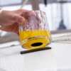 Hand holding a faceted glass with yellow liquid, glass tilted over a small black rectangular object on a light ribbed surface; blurred window background