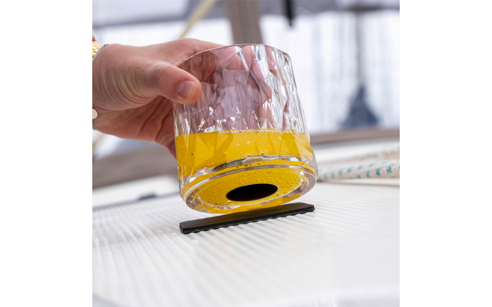Hand holding a faceted glass with yellow liquid, glass tilted over a small black rectangular object on a light ribbed surface; blurred window background