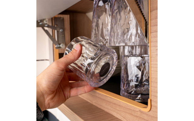 Hand holding a faceted tumbler in front of a wooden compartment; inside are two other faceted glasses and a decanter; metal hinge visible