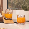 Two cut-glass tumblers with orange-colored liquid on a kitchen countertop; one tumbler leans against a small wooden board; window with trees in the background; a dish brush at left