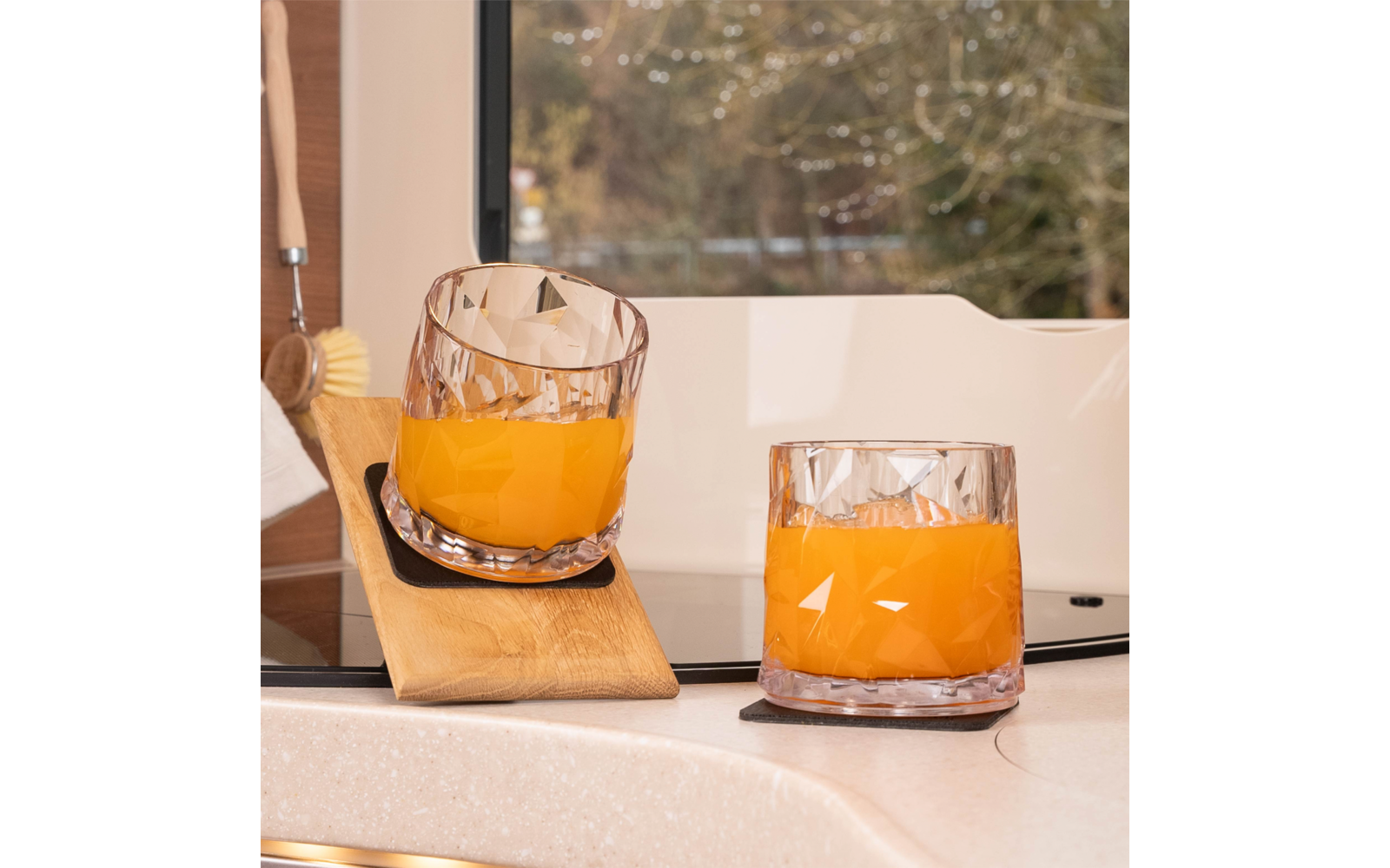 Two cut-glass tumblers with orange-colored liquid on a kitchen countertop; one tumbler leans against a small wooden board; window with trees in the background; a dish brush at left