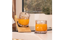 Deux verres taillés contenant un liquide orange sur un plan de travail de cuisine; un verre appuyé contre une petite planche en bois; fenêtre avec des arbres en arrière-plan; à gauche une brosse de vaisselle