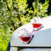 Two faceted glasses with red liquid on the hood of a white car, green foliage in the background