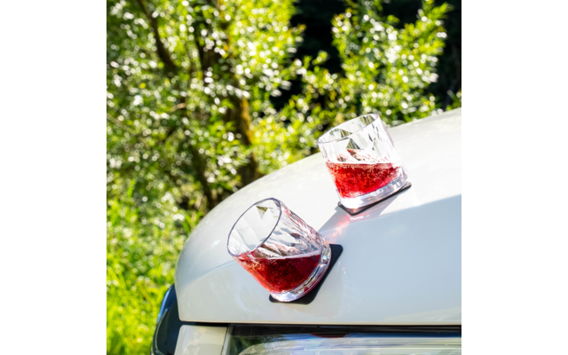 Two faceted glasses with red liquid on the hood of a white car, green foliage in the background