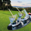 Two clear glasses with ice, mint leaves, blueberries and striped paper straws on a black tray held by a hand; green lawn, trees, hills and sky in the background