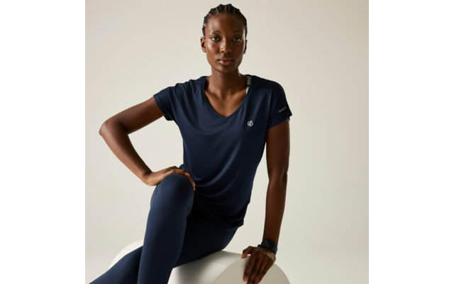 Person sitzt auf rundem weißen Hocker, trägt dunkelblaues V‑Shirt und dunkelblaue Leggings, geflochtene Haare, rechte Hand auf dem Knie, linke Hand mit Uhr auf dem Hocker, heller einfarbiger Hintergrund