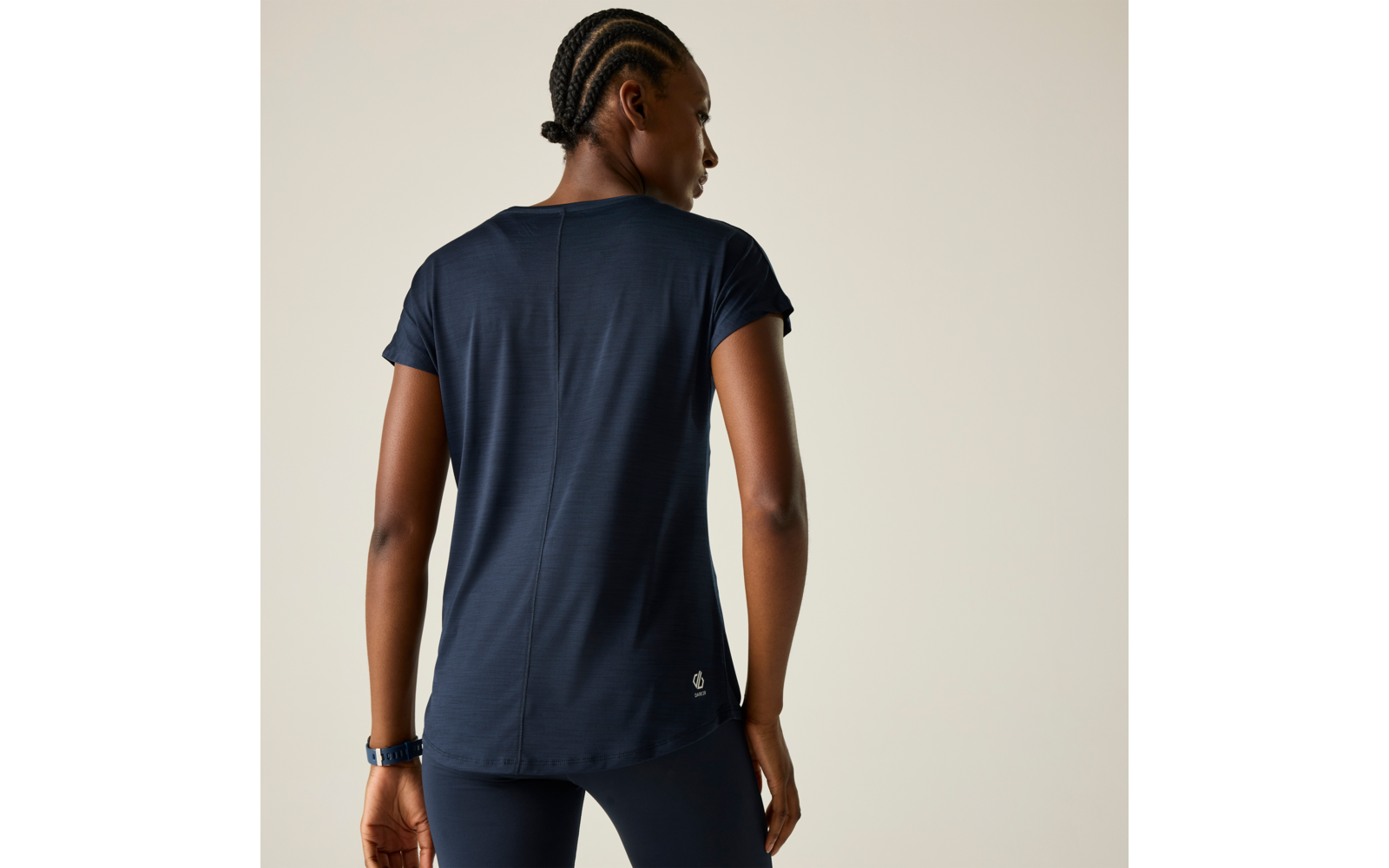 Person von hinten, trägt dunkelblaues kurzärmeliges Shirt und passende Leggings, geflochtene Haare am Nacken gebunden, Uhr am linken Handgelenk, heller beiger Hintergrund, kleines Logo am unteren Rückensaum